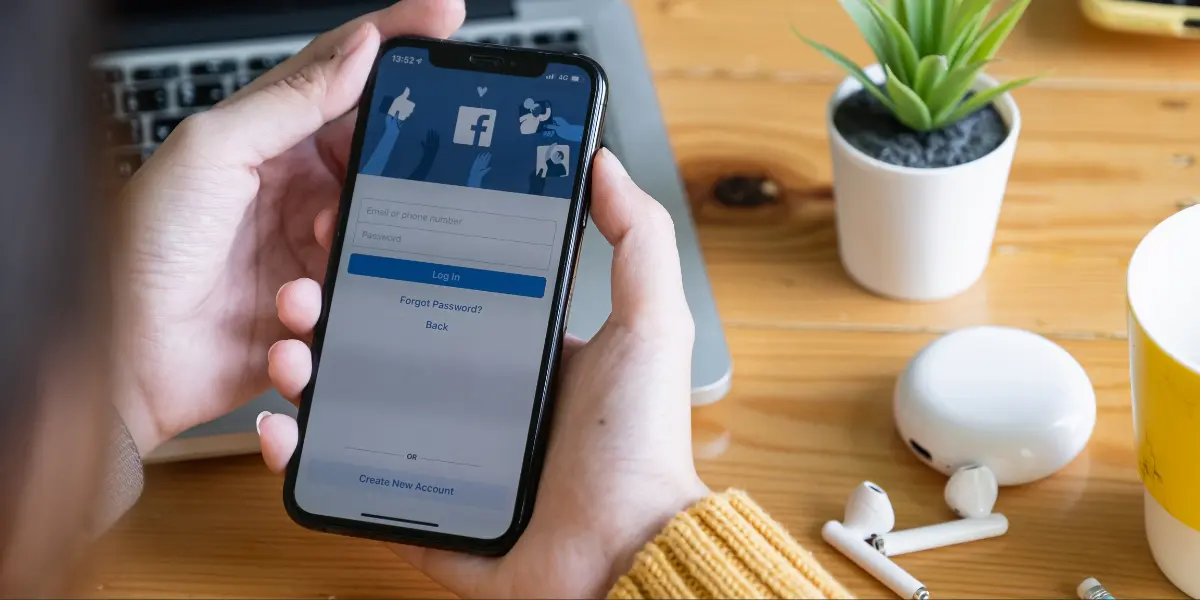 A woman in a yellow jumper holding a smartphone that's showing the Facebook log in page