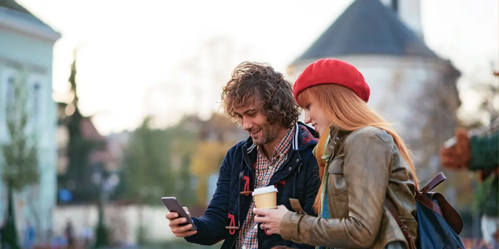 A man and a woman looking at a phone being held by the man, whilst the woman holds a cup of coffee in an outdoor setting