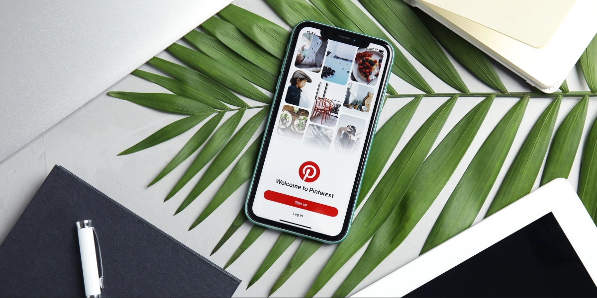 photo shows a phone showing the marketing on pinterest log in screen, resting on a table with green leaves and a notebook